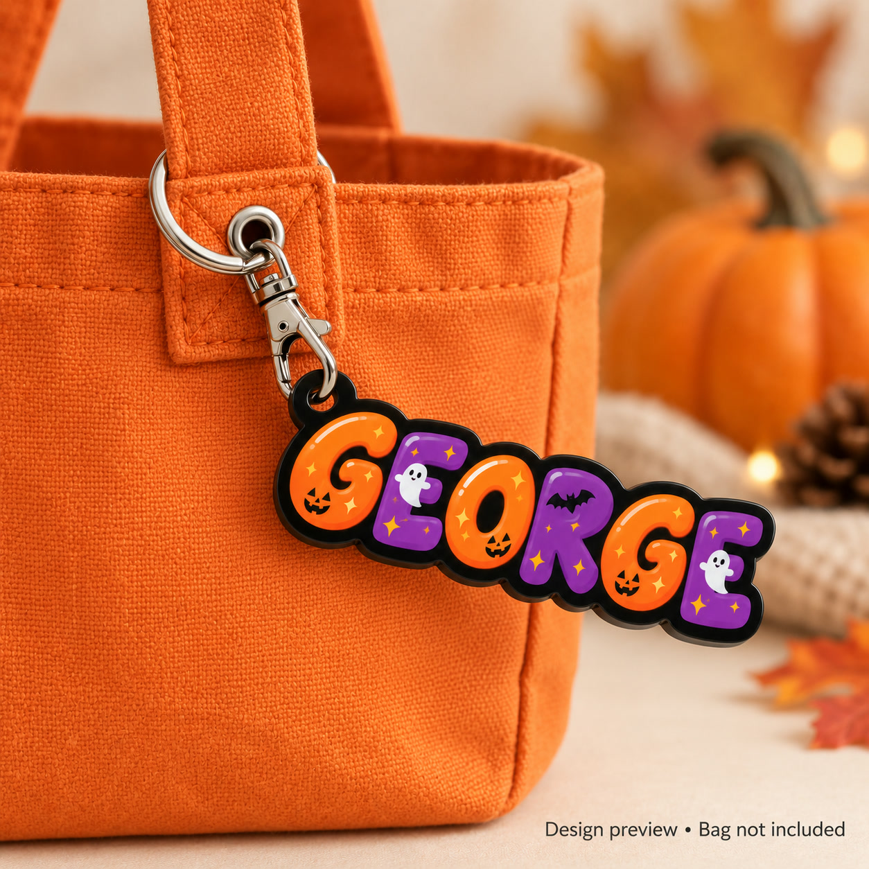 Lifestyle design preview of the GEORGE Halloween bubble-name charm on black acrylic hanging from an orange trick-or-treat bag; silver lobster clasp faces into the charm. Bag not included.