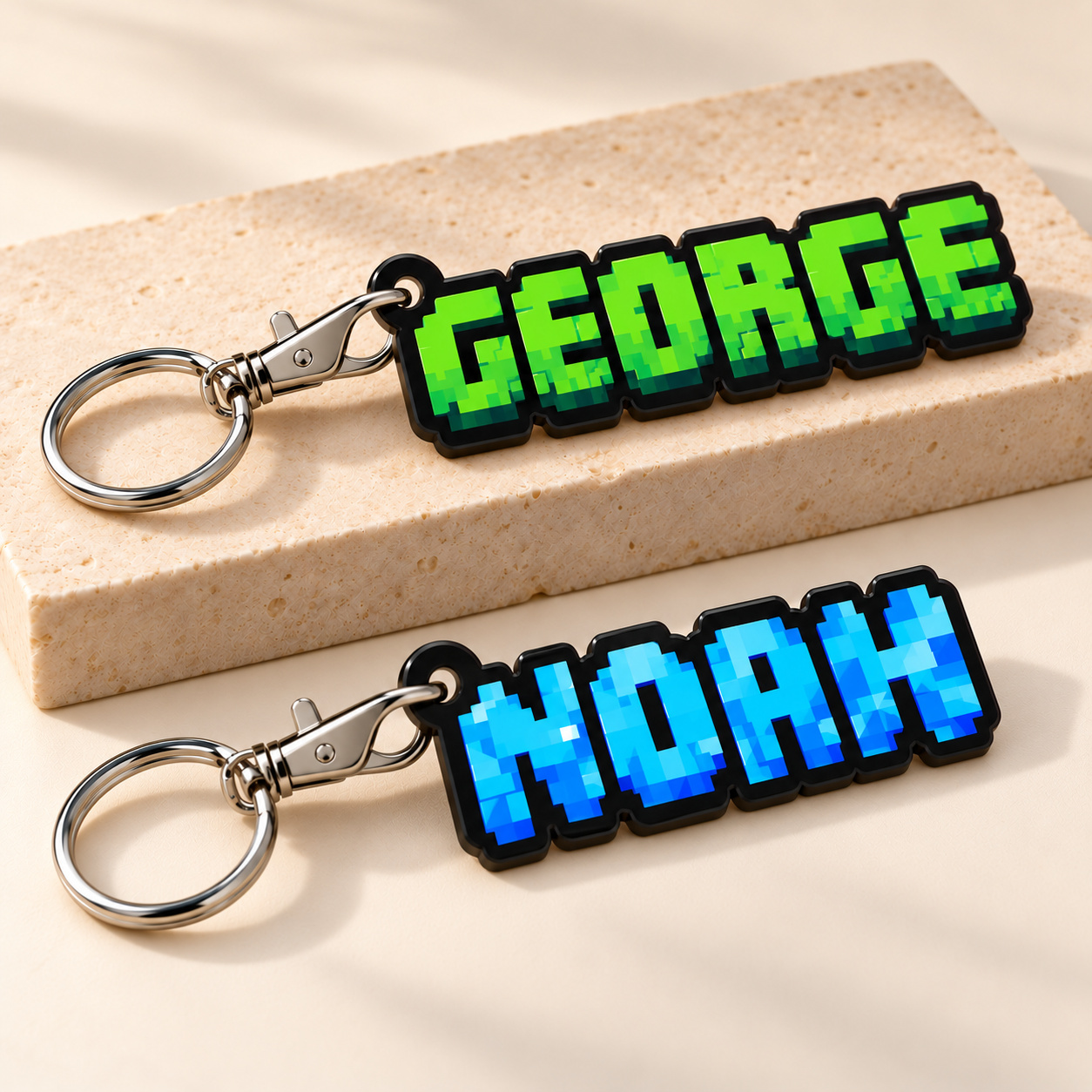 Studio digital mockup of GEORGE in grass-green and NOAH in crystal-blue pixel lettering on name-shaped black acrylic keyrings, each with an inward silver lobster clasp and outward round split ring.