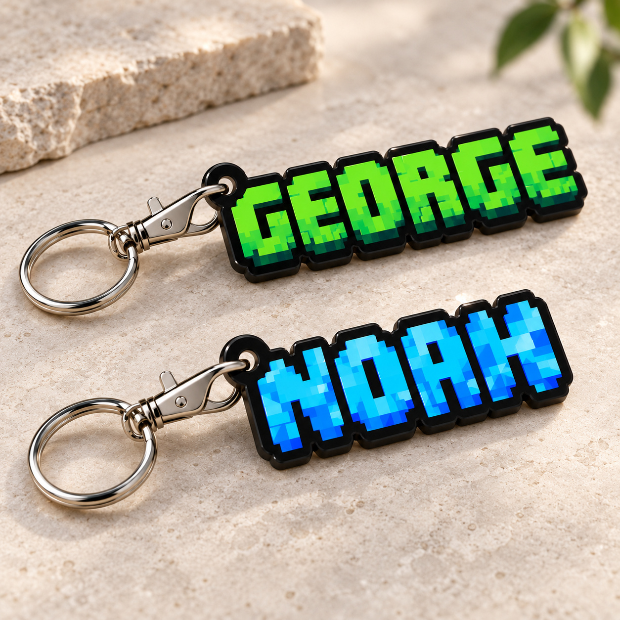 Close-up digital mockup of GEORGE in green and NOAH in blue pixel lettering, UV printed on contour-cut black acrylic keyrings with silver lobster clasps and split rings.